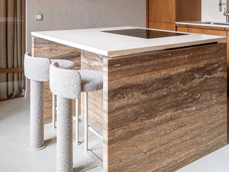May include: A modern kitchen island with a white countertop and a built-in cooktop. The island base is made of brown, textured stone. Two white, upholstered bar stools with curved backs and white metal legs are positioned in front of the island.