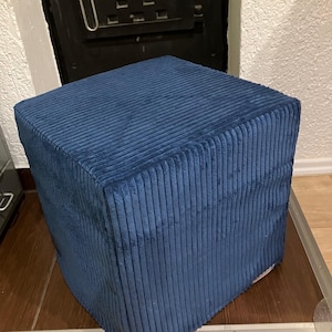 Può includere: Un pouf quadrato in velluto a coste blu navy. Il tessuto ha una trama a coste verticali. Il pouf è posto su un pavimento in legno. Lo sfondo comprende una porta in metallo nero e una parete bianca.