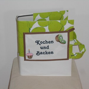 Può includere: Un raccoglitore in tessuto verde e bianco con una toppa ricamata marrone che dice "Kochen und Backen" (tedesco per "Cucinare e cuocere"). Il raccoglitore ha un design a kiwi e un piccolo cupcake con glassa rosa e una candela.