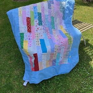 Op de afbeelding: Een kleurrijke patchworkquilt met een lichtblauwe rand ligt op een houten bank en gras. De quilt heeft verticale stroken van verschillende stoffen met patronen in roze, blauw, groen en geel. De quilt is ongeveer 178 cm lang en 152 cm breed.