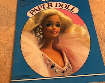 May include: A vintage Barbie paper doll book with a pre-cut doll and fashions. The cover features a colorful illustration of Barbie wearing a pink and white outfit with silver stars.