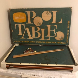 Może przedstawiać: Zestaw do gry w bilard stołowy vintage marki Tudor z zielonym stołem pokrytym suknem, dwoma drewnianymi kijami i kompletem bil. Na pudełku widnieje napis "Pool Table" dużymi żółtymi literami na zielonym tle. Pudełko zawiera również napis "Fun for the entire family - Sturdy Tru-Scale Table Top Model - Automatic Ball Return."