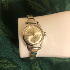 Puede incluir: Un reloj de pulsera plateado con una esfera blanca y una correa plateada. La esfera del reloj tiene marcas negras y una ventana de fecha en la posición de las 3 en punto. La esfera del reloj también tiene el texto "Rostone Automatic 17 Jewels" impreso en ella.