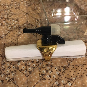 May include: A gold ring with a black plastic gun attached to the top. The gun has a white plastic tip and a small black handle.