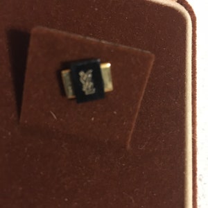 Pode incluir: Uma pequena caixa retangular preta com uma moldura dourada. A caixa tem um logotipo na parte superior que &eacute; uma letra Y estilizada.
