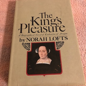 May include: A tan book cover with the title "The King's Pleasure" in black text. The subtitle is "A Biographical Novel of Katharine of Aragon" in smaller black text. The author's name is "by NORAH LOFTS" in black text. A portrait of a woman in a black dress with a white collar is in a red frame on the cover.