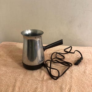 Pode incluir: Uma cafeteira elétrica com corpo de metal prateado polido, base de plástico preto e alça de plástico preto. O aparelho possui um longo cabo de alimentação preto com plugue, que se estende a partir da lateral da alça.