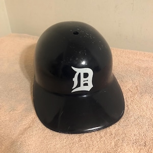 May include: Black baseball helmet with a white Detroit Tigers logo. The helmet has a black visor.