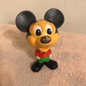 Op de afbeelding: Een vintage Mickey Mouse figuur van plastic in de kleuren geel, zwart, rood en groen. Het figuur staat met zijn armen langs zijn zijde en heeft een brede glimlach.