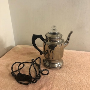 Puede incluir: Una cafetera eléctrica cromada vintage con un asa negra y una tapa de cristal. La cafetera tiene un pico y está sobre una superficie rosa. Un cable en blanco y negro está enrollado cerca.