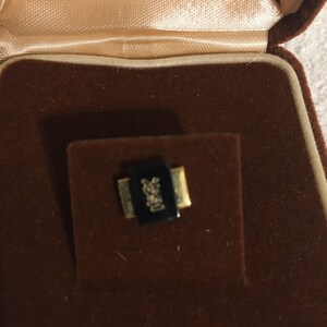 Pode incluir: Um broche com o logotipo YSL preto e dourado em uma caixa de veludo marrom.