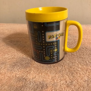 May include: A yellow and black Pac-Man mug with a Pac-Man maze design. The mug has a yellow handle and a yellow rim.