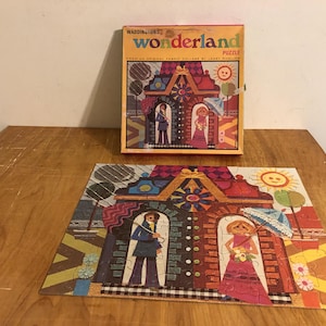 May include: Vintage Waddington's Wonderland puzzle with a colorful illustration of a building with figures. The puzzle is partially assembled on a wooden surface, with the box nearby. The box features the puzzle's title and artwork.