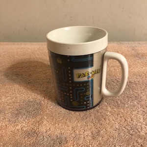 May include: White ceramic mug with a black, blue, and yellow Pac-Man design. The mug has a handle and says "Pac-Man" on the side.