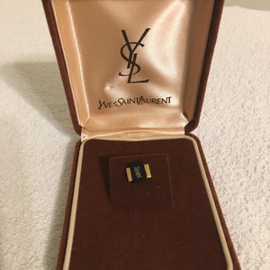 Pode incluir: Uma caixa de joias de veludo marrom com um interior de cor creme. A caixa tem o logotipo Yves Saint Laurent no interior da tampa. A caixa est&aacute; aberta e cont&eacute;m uma pequena joia dourada.