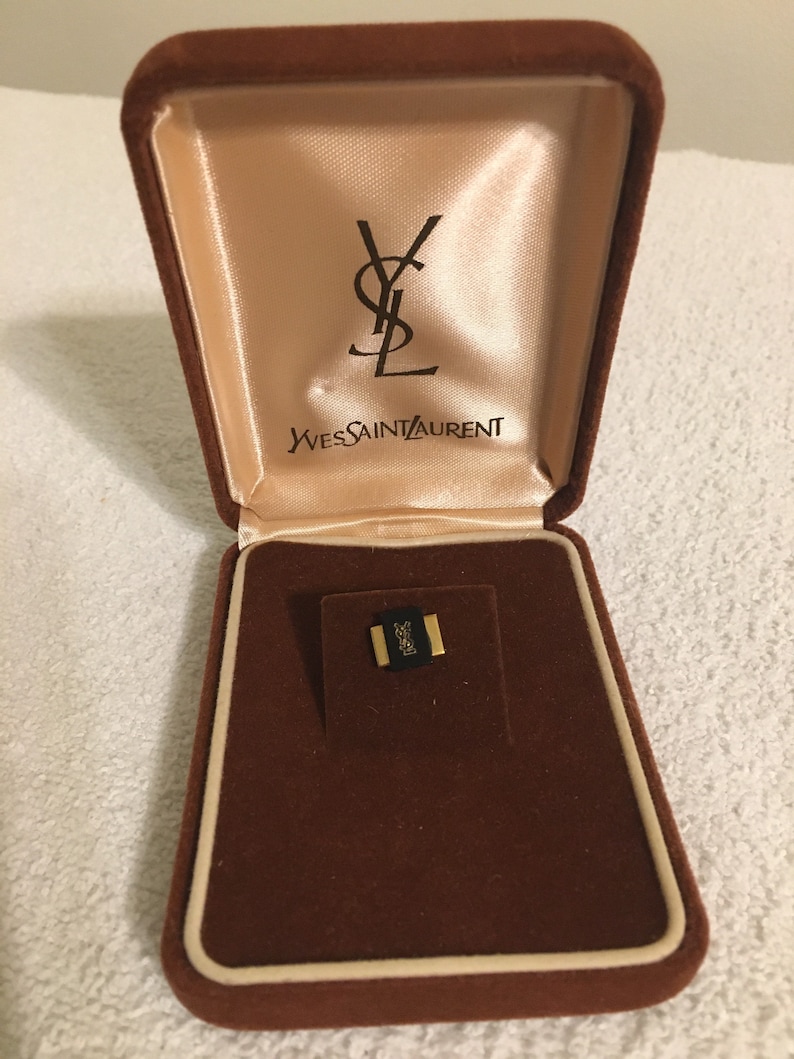Pode incluir: Uma caixa de joias de veludo marrom com um interior de cor creme. A caixa tem o logotipo Yves Saint Laurent no interior da tampa. A caixa est&aacute; aberta e cont&eacute;m uma pequena joia dourada.
