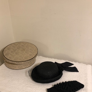 May include: A black hat with a wide brim and a black bow. The hat is sitting on a white surface next to a black knitted headband.