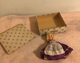 Puede incluir: Una muñeca vintage con cabello rubio que lleva un vestido morado con una sobrecapa de encaje blanco y un patrón floral. La muñeca está en una caja marrón con lunares blancos.