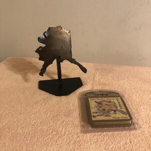 May include: A metal sculpture of the state of Alaska on a black stand, next to a set of Alaskan coasters. The coasters feature a map of Alaska and are packaged in clear plastic. The sculpture is made of dark metal.