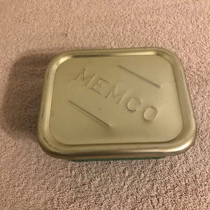 Könnte beinhalten: Ein rechteckiger Metallbehälter mit dem Wort "MEMCO" auf dem Deckel. Der Deckel hat eine helle, gedämpfte Goldfarbe mit leicht abgerundeten Kanten. Der Behälter scheint auf einer strukturierten Oberfläche zu liegen.