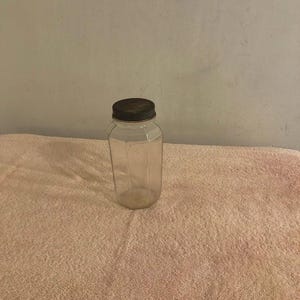 Pode incluir: Um frasco de vidro transparente com corpo hexagonal e uma tampa de rosca de metal escuro e texturizado. O recipiente apresenta seis painéis verticais que se encontram em bordas definidas, criando uma forma geométrica.
