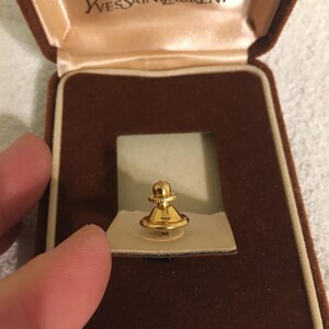 Pode incluir: Um prendedor de gravata dourado em uma caixa de veludo marrom com as palavras "Yves Saint Laurent" na tampa.