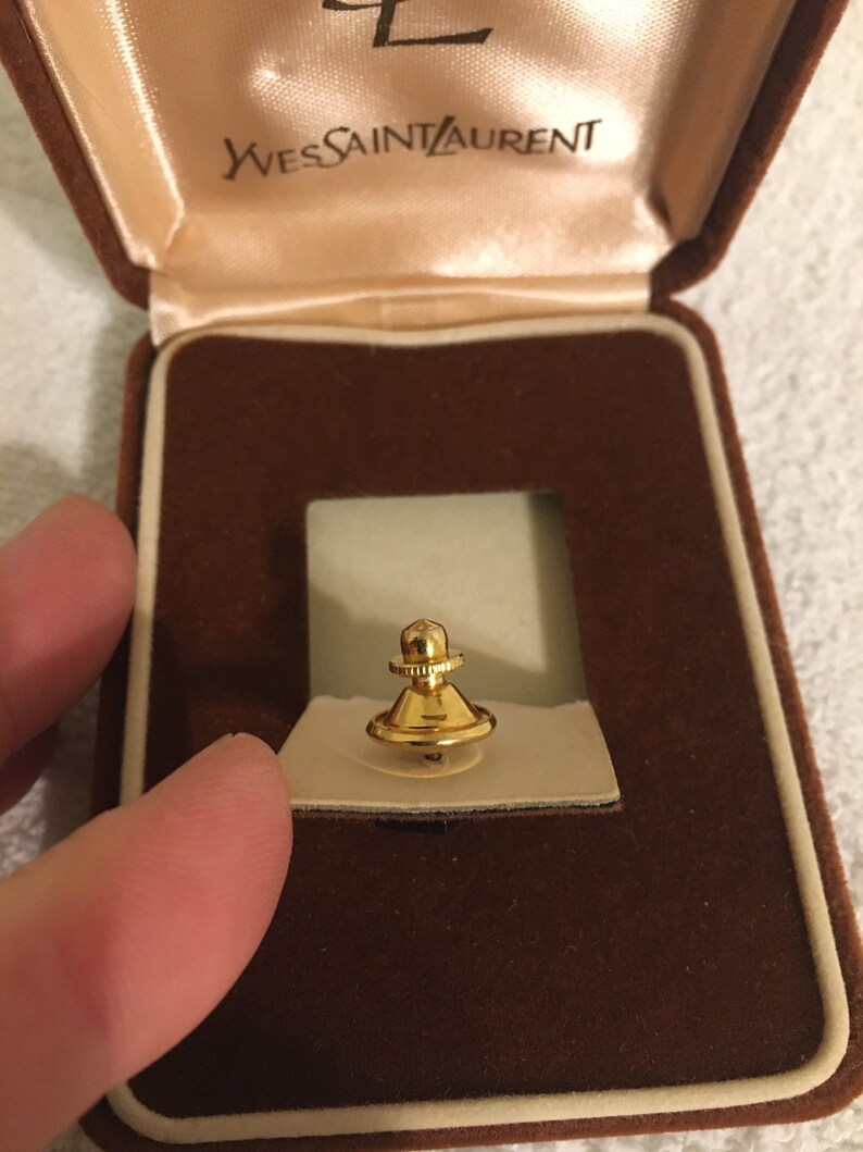 Pode incluir: Um prendedor de gravata dourado em uma caixa de veludo marrom com as palavras "Yves Saint Laurent" na tampa.
