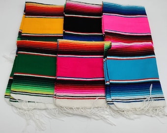 May include: A collection of colorful serape blankets, each folded and displaying a distinct color scheme. The blankets feature horizontal stripes in various hues, including yellow, black, pink, green, and blue, with white fringe along the edges. These blankets are a vibrant representation of Mexican culture.