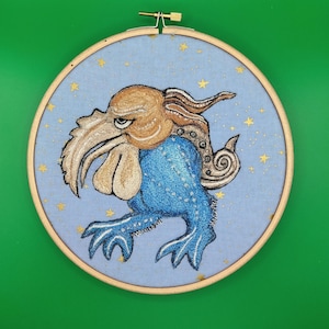 May include: Embroidered artwork in a wooden hoop. The design features a blue creature with a large beak, a tan head, and blue body. The background is light blue with gold star accents. The artwork is set against a green background.