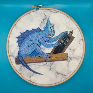 May include: Embroidered artwork in a wooden hoop. A blue creature with a finned tail and spiky head is writing on a chalkboard that reads "FAKE NEWS". The background is white with a marble pattern.
