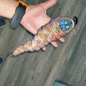 May include: Embroidered caterpillar plush toy with a brown body, colorful circular markings, and a blue face. The caterpillar has gold antennae and a black loop for hanging. The plush is held in a person's hand.