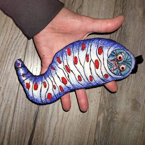 May include: A blue and white embroidered patch of a cartoon worm with red spots and a scary face. The worm is curled up and has a black strap on the back.