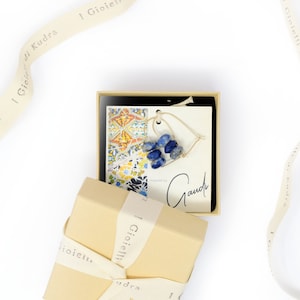 Peut inclure: Une petite boîte cadeau nouée d'un ruban blanc avec le texte "I Gioielli di Kudra" imprimé dessus. La boîte est ouverte et à l'intérieur se trouve une petite boîte noire avec un collier en forme de cœur bleu et blanc à l'intérieur. Le collier est sur une carte blanche avec le texte "Gauche" écrit dessus.