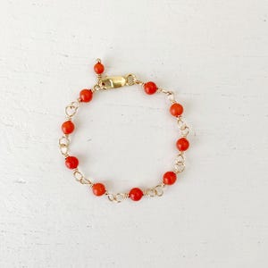 Peut inclure: Un bracelet doré orné de perles rouges vives. Les perles sont enfilées sur un fil d'or fin, créant un motif de chaîne. Le bracelet est doté d'un fermoir sécurisé et d'une petite perle pendante. Le bijou est présenté sur une surface blanche. Mesure environ 18 cm.