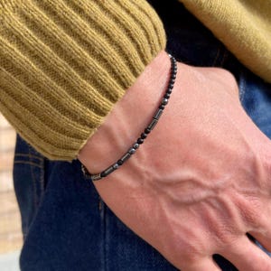 Peut inclure: Un bracelet de perles composé de petites perles noires et d'éléments cylindriques et sphériques argentés. Le bracelet est porté sur un poignet, avec un pull vert et un jean bleu en arrière-plan.