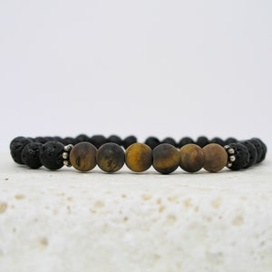 Puede incluir: Un brazalete de cuentas de lava negra y ojo de tigre. El brazalete tiene cuentas espaciadoras plateadas.