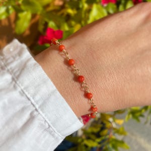 Puede incluir: Una delicada pulsera dorada con pequeñas cuentas redondas de color naranja. La pulsera se lleva en la muñeca, con una camisa blanca visible en el fondo. El diseño es sencillo y elegante.