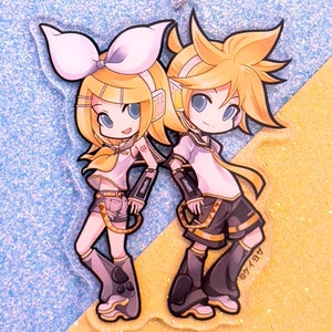 May include: A clear acrylic keychain featuring two anime characters, a girl with a pink bow and a boy with yellow hair. The characters are standing side-by-side and are holding hands. The keychain has a silver chain attached to it. The text "©ケイコ" is printed on the bottom right corner of the keychain.