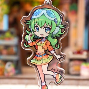 May include: A clear acrylic keychain depicting a colourful anime-style character with green hair, blue goggles, and an orange jacket. The character is smiling and wearing a yellow top and skirt. The keychain is attached to a silver chain.