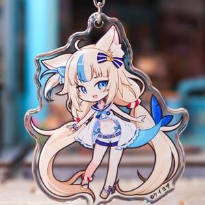 May include: A clear acrylic keychain featuring a cartoon character with long blonde hair, cat-like ears, and a blue shark tail. The character wears a white and blue sailor-themed outfit with a bow. The text "ケイヨザ" is printed in the bottom right corner.