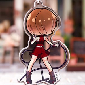 May include: A clear acrylic keychain featuring a cartoon illustration of a character with short brown hair, wearing a red sleeveless top and a matching red skirt. The character stands at a microphone stand, holding a microphone, with a black cable.