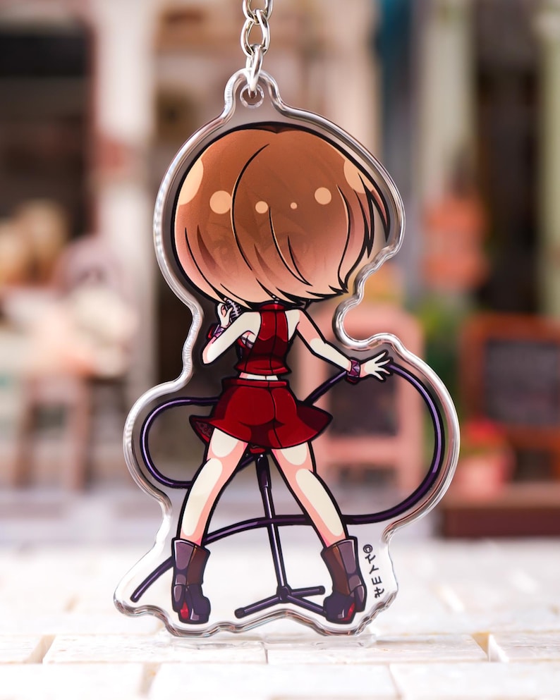 May include: A clear acrylic keychain featuring a cartoon illustration of a character with short brown hair, wearing a red sleeveless top and a matching red skirt. The character stands at a microphone stand, holding a microphone, with a black cable.
