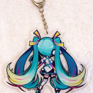 Peut inclure: Un porte-cl&eacute;s repr&eacute;sentant un personnage d'anime turquoise et jaune avec de longs cheveux bleus et une tenue blanche. Le porte-cl&eacute;s est attach&eacute; &agrave; une cha&icirc;ne argent&eacute;e et &agrave; un anneau argent&eacute;.