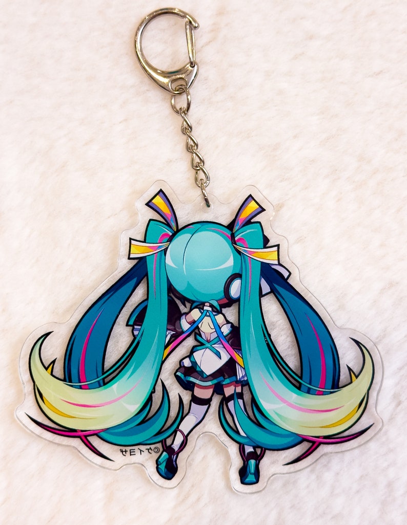 Peut inclure: Un porte-cl&eacute;s repr&eacute;sentant un personnage d'anime turquoise et jaune avec de longs cheveux bleus et une tenue blanche. Le porte-cl&eacute;s est attach&eacute; &agrave; une cha&icirc;ne argent&eacute;e et &agrave; un anneau argent&eacute;.