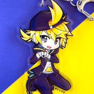 May include: A yellow-haired anime character with a blue and black outfit, wearing a black baseball cap, is kneeling with their hands in a heart shape. The character is on a clear acrylic keychain with a silver chain.