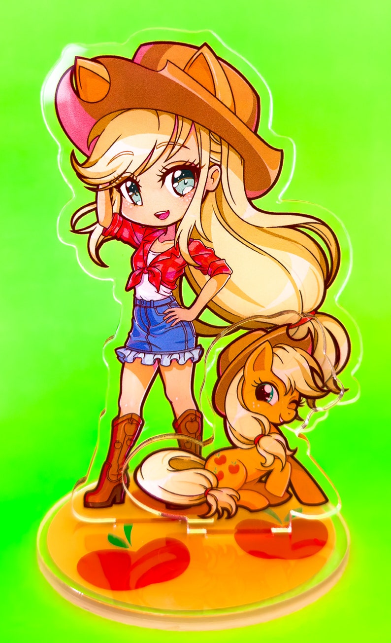 May include: A clear acrylic stand with a cartoon image of a female character wearing a red shirt, blue jeans, and a cowboy hat. The character is standing with her right hand on her hip. A smaller cartoon image of a pony is sitting at her feet. The stand has a yellow base with red apples.