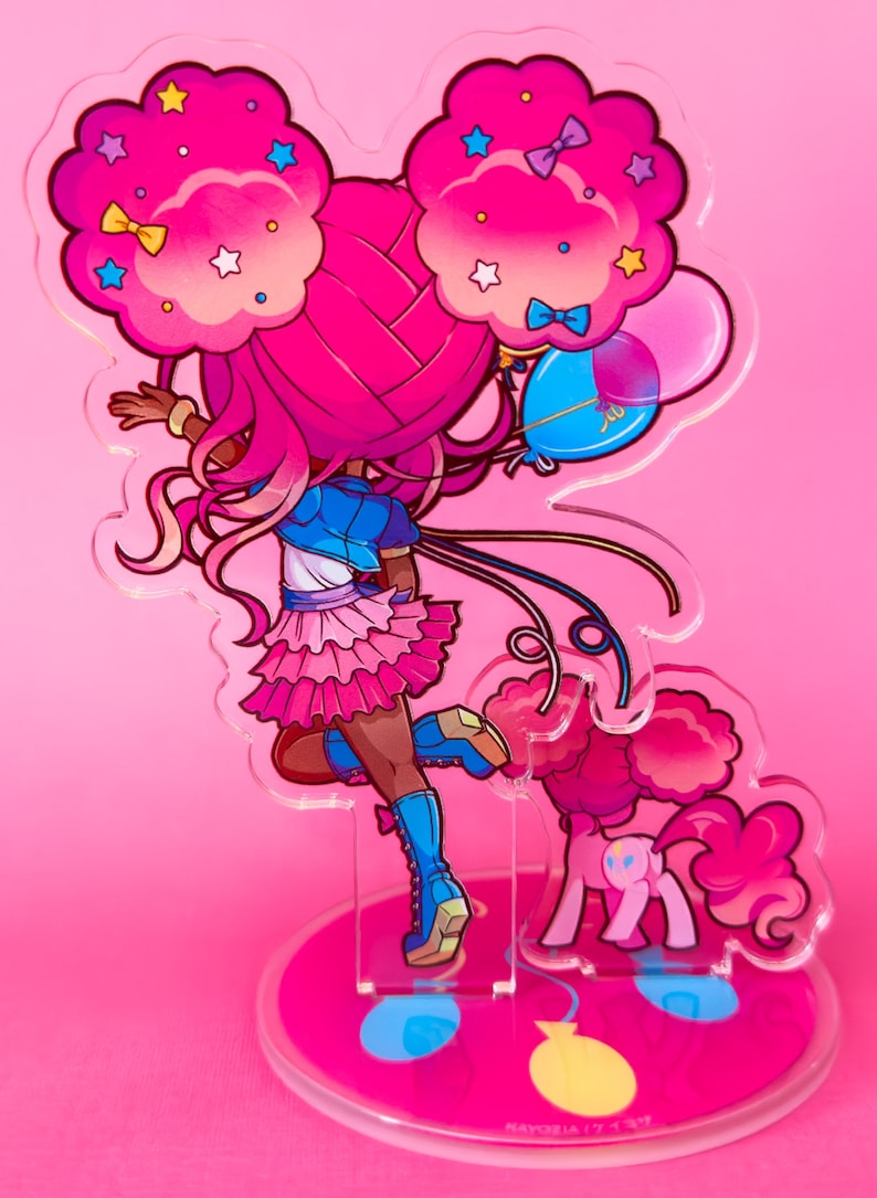 Puede incluir: Un personaje rosa y azul con grandes lazos rosas en el pelo y una falda rosa que sostiene globos. El personaje est&aacute; de pie sobre una base redonda con un dise&ntilde;o rosa y azul. La base tiene el texto "ましろ屋" en japon&eacute;s.