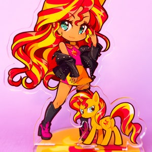 Puede incluir: Un soporte de acr&iacute;lico con una imagen de dibujos animados de un pony con una melena y cola amarillas. El pony lleva una camisa rosa y una chaqueta de cuero negra. El pony est&aacute; de pie sobre un dise&ntilde;o de sol amarillo y rojo.