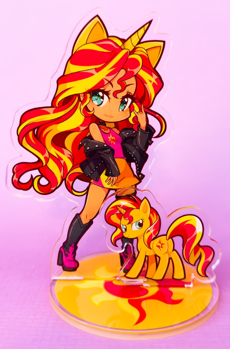 Puede incluir: Un soporte de acr&iacute;lico con una imagen de dibujos animados de un pony con una melena y cola amarillas. El pony lleva una camisa rosa y una chaqueta de cuero negra. El pony est&aacute; de pie sobre un dise&ntilde;o de sol amarillo y rojo.