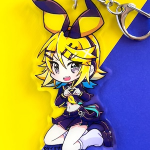 May include: A yellow and black anime character keychain with a heart-shaped hand gesture. The character has long yellow hair, blue eyes, and is wearing a black and blue outfit. The keychain is on a yellow and blue background.